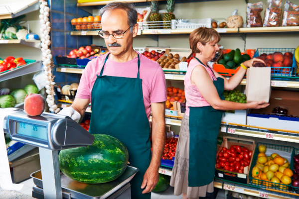 Dona de mercadinho independente atrás do balcão, com gôndola abastecida de arroz, feijão e óleo ao fundo