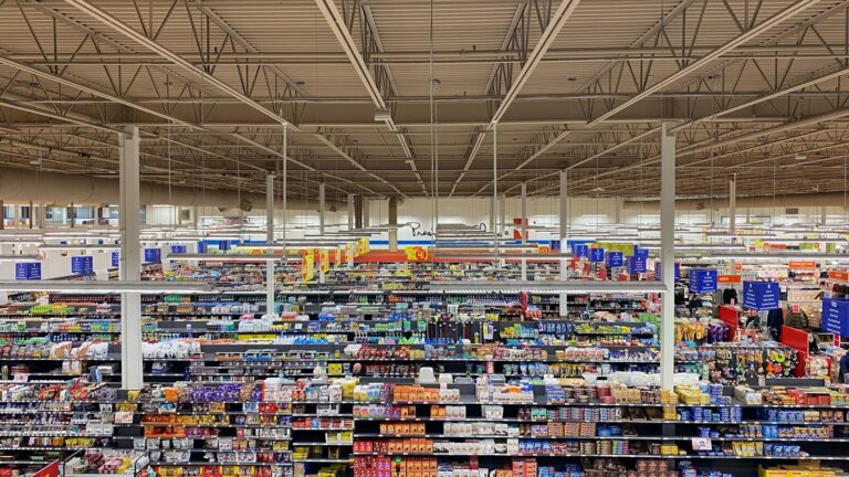 Gôndola de supermercado de bairro com etiquetas de preço terminando em ,99 e calculadora ao lado